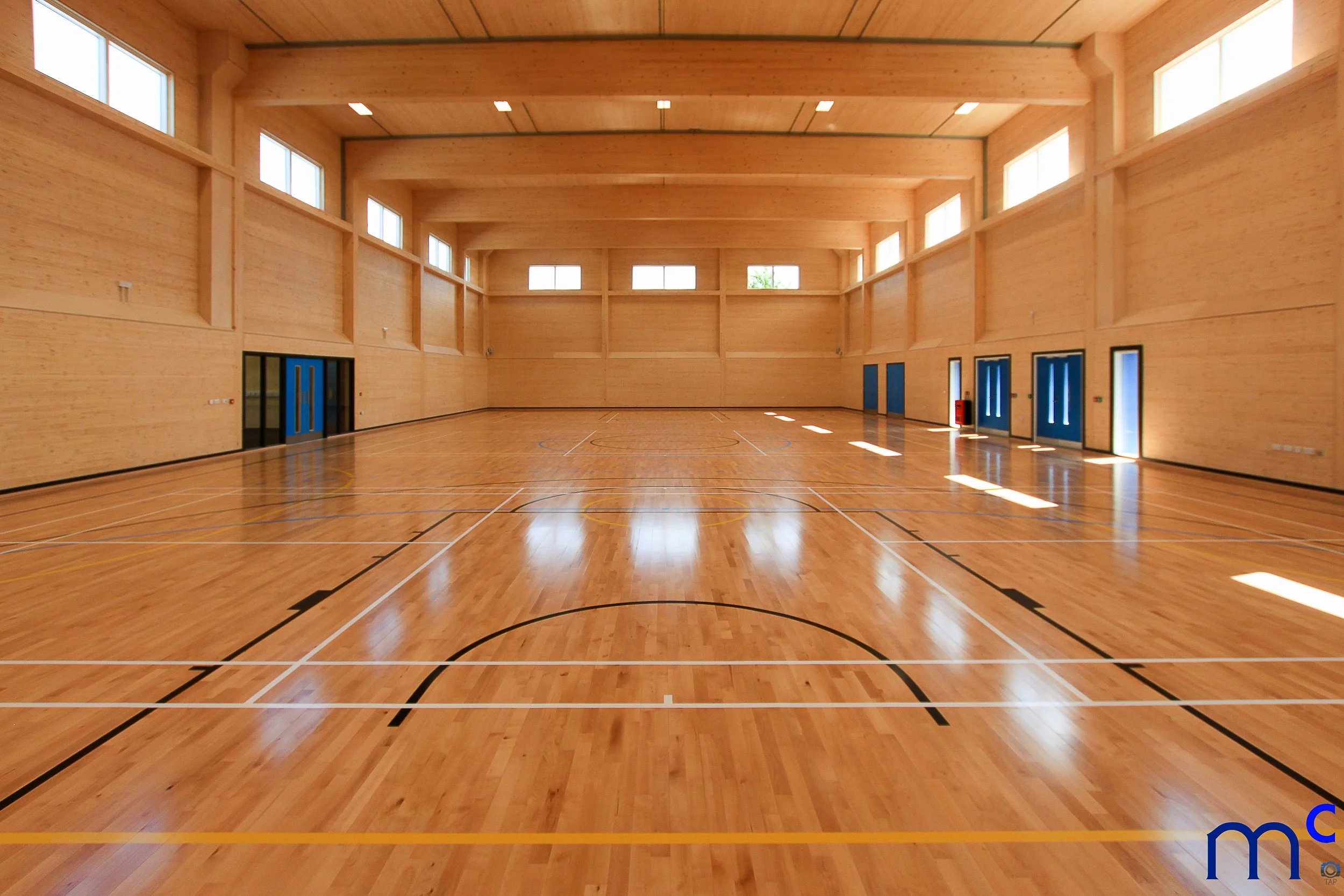 An empty indoor gymnasium with a polished wooden floor, light wood-paneled walls, and multiple windows allowing natural light. Blue double doors are along the walls, and lines for sports are marked on the floor.