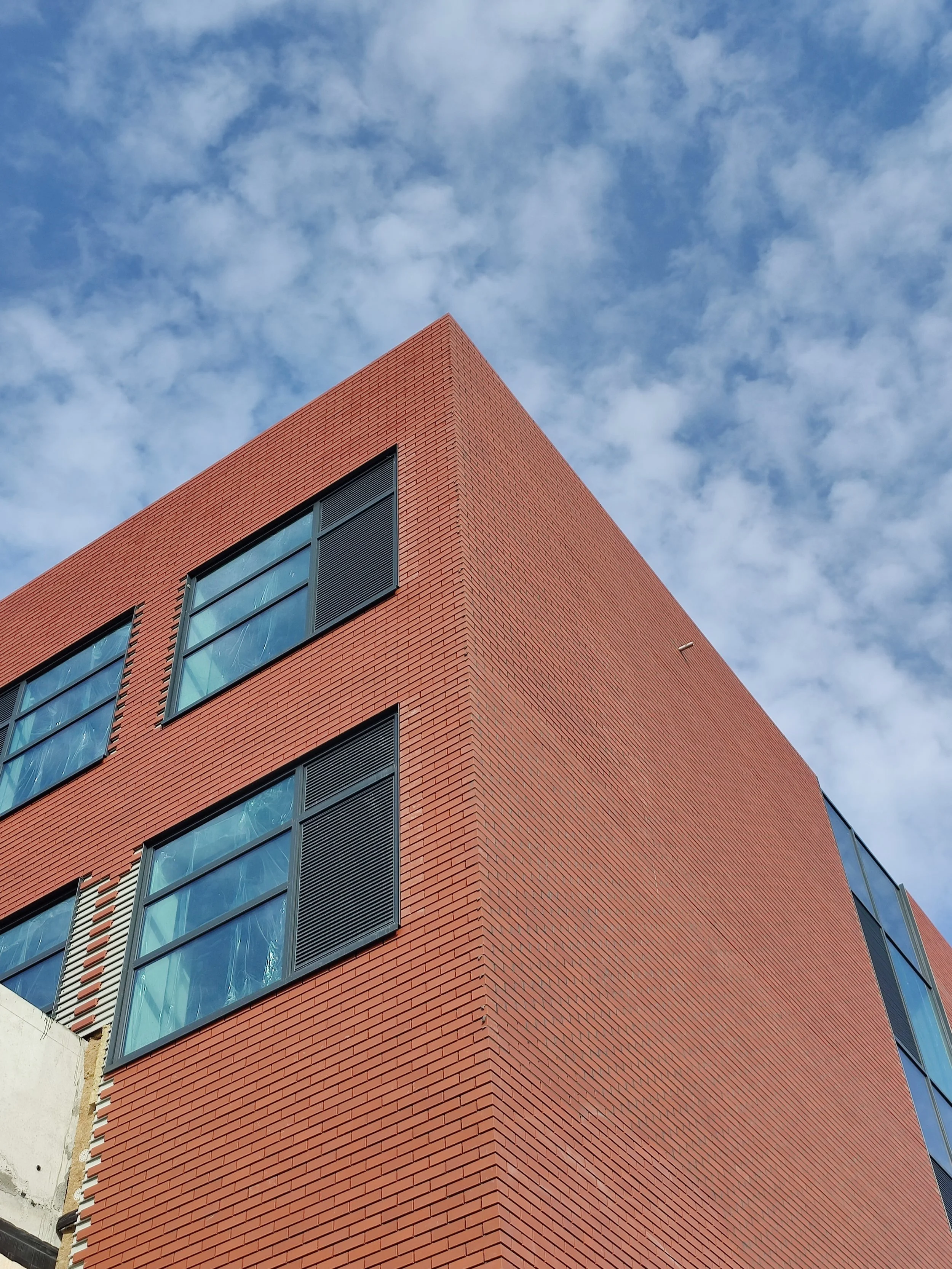 A modern red brick building with large black-framed windows set against a blue sky with scattered clouds.