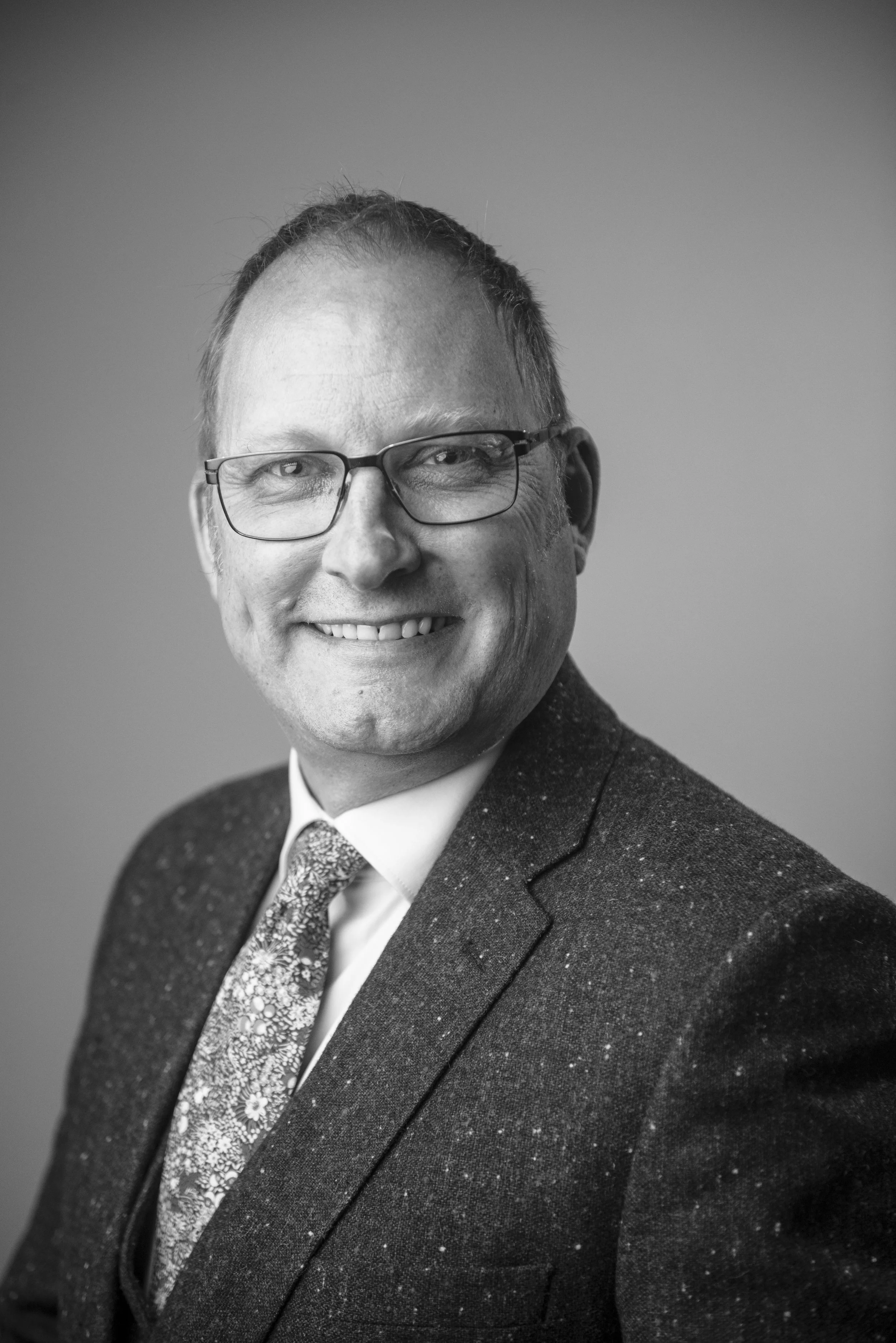Black and white portrait of a man smiling, wearing glasses, a suit, and a patterned tie.