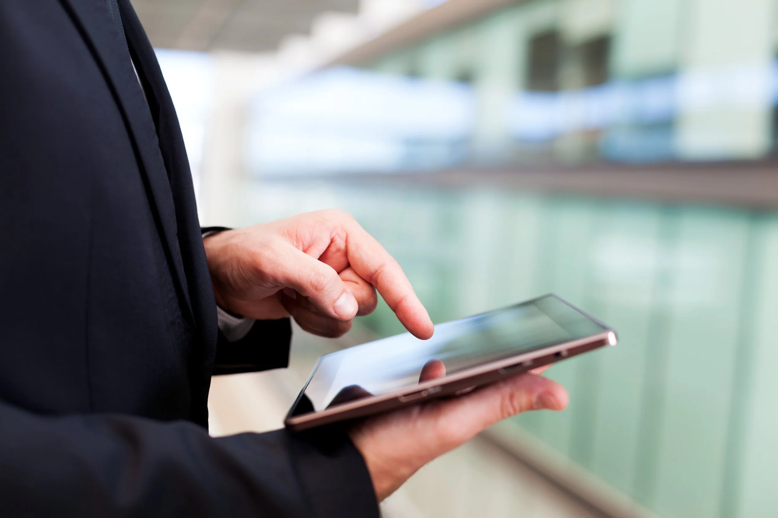 Person in a dark suit using a smartphone with blurred modern architecture in the background.