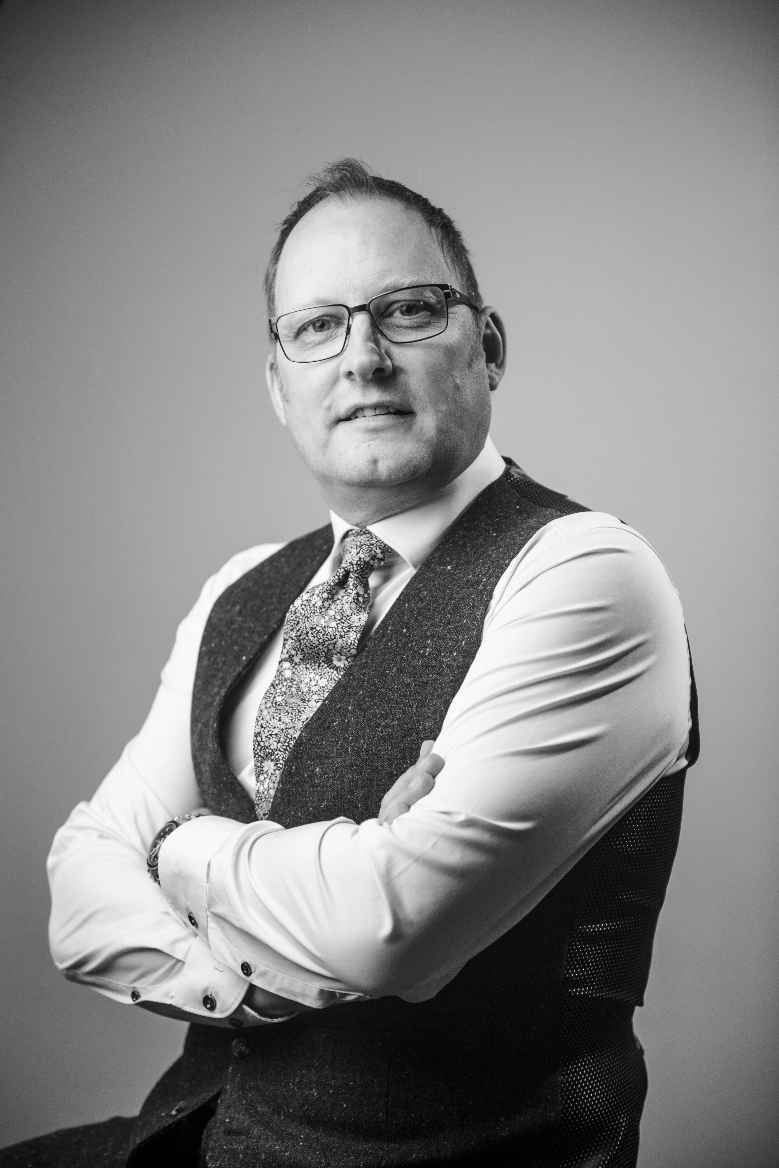 Black and white portrait of a man with glasses, wearing a vest over a white shirt with rolled-up sleeves, and a patterned tie, standing with arms crossed against a plain background.
