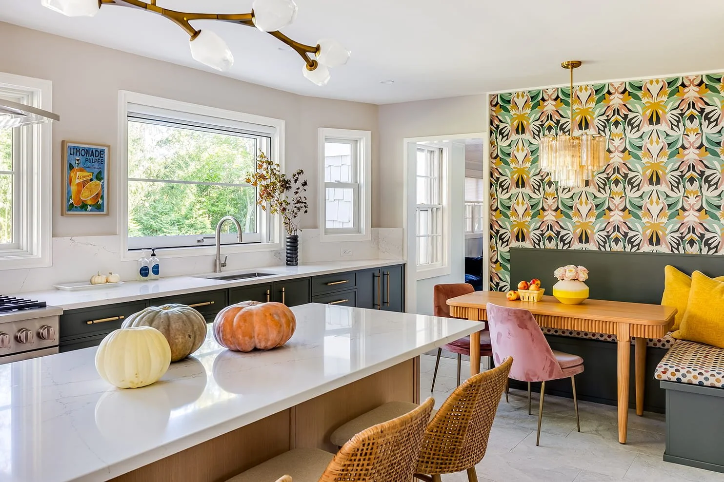 Modern kitchen with white countertops, dark green cabinets, and a colorful floral accent wall. Fresh pumpkins on the island, a light fixture above the table, and a window view of greenery outside.