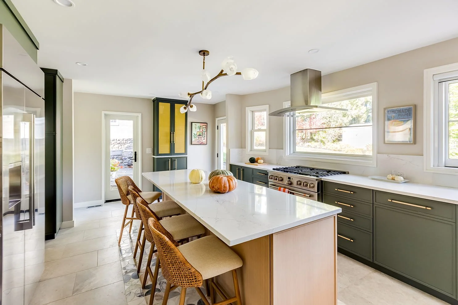 Modern kitchen with green cabinets, white countertops, and a large island with four barstools, decorative pumpkins, and a stainless steel range hood.