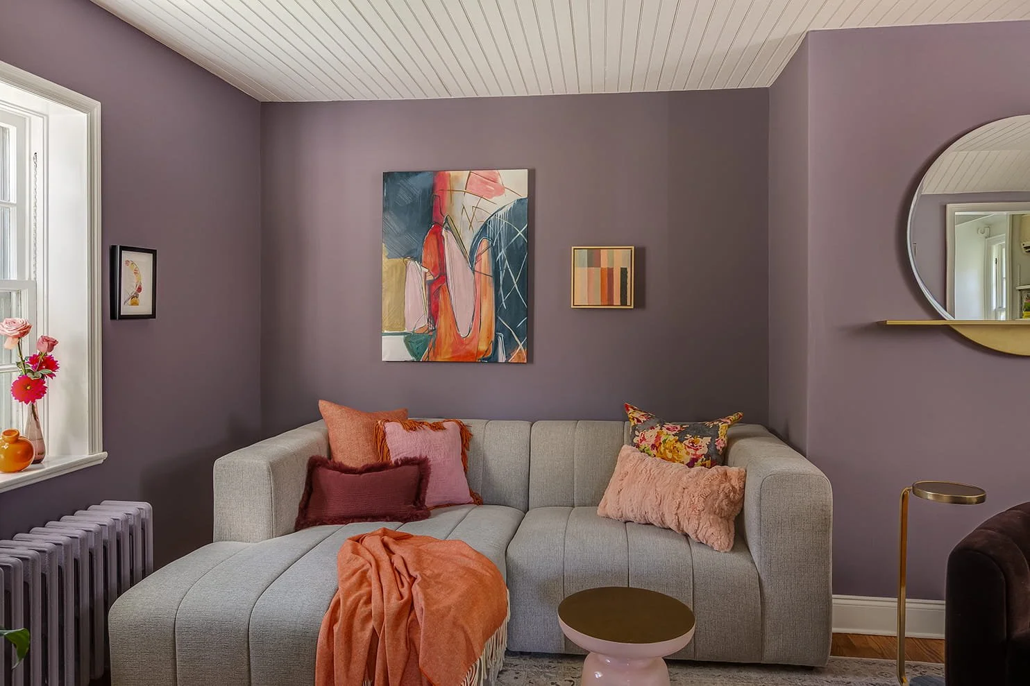 Living room with purple walls, beige sofa with pink and orange cushions, abstract wall art, round mirror, window with pink flowers, orange blanket, gold-accented side table and lamp.