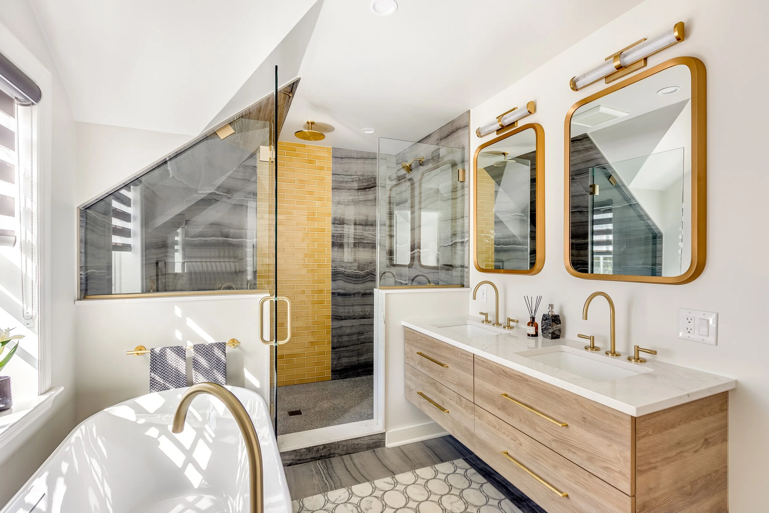 Modern bathroom with dual gold faucets, wooden vanity with gold handles, large mirrors with gold frames, glass shower with yellow brick and grey marble walls, white bathtub, window with blinds, and decorative items on the counter.