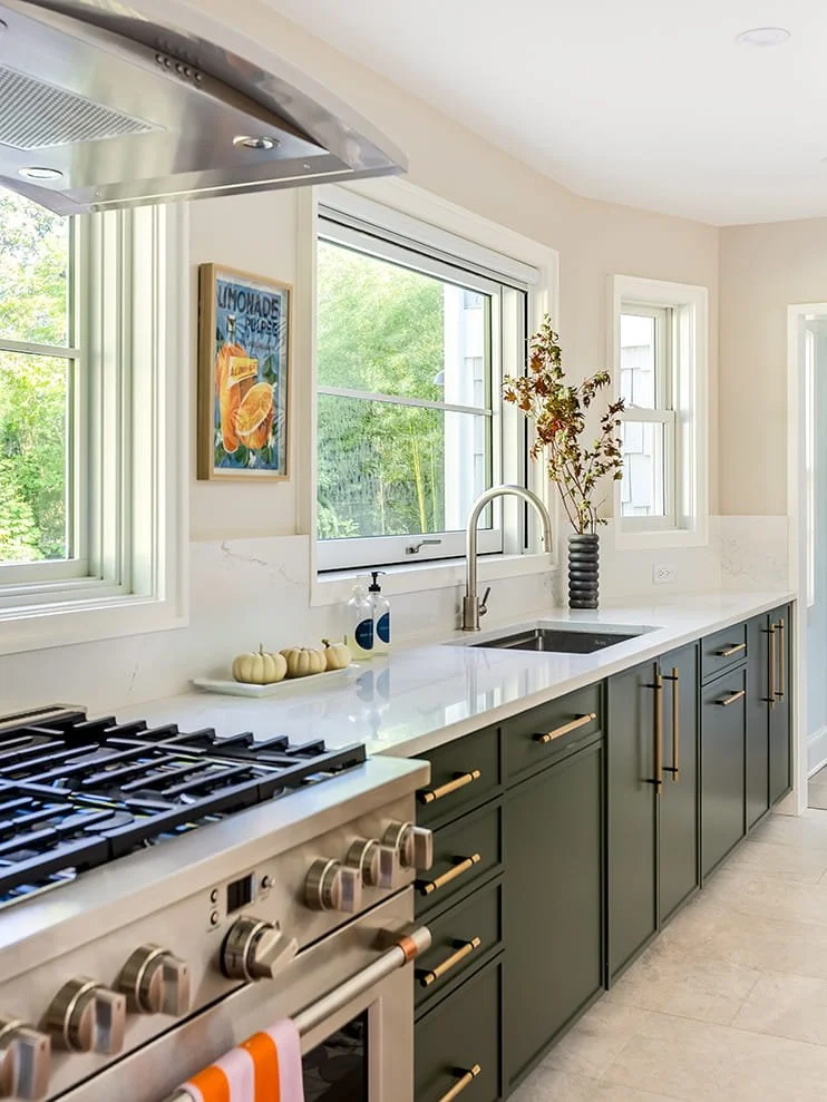 Modern kitchen with white countertops, dark green cabinets with gold handles, stainless steel stove and range hood, white marble backsplash, large windows, a vase with branches, and a wall poster of lemonade.