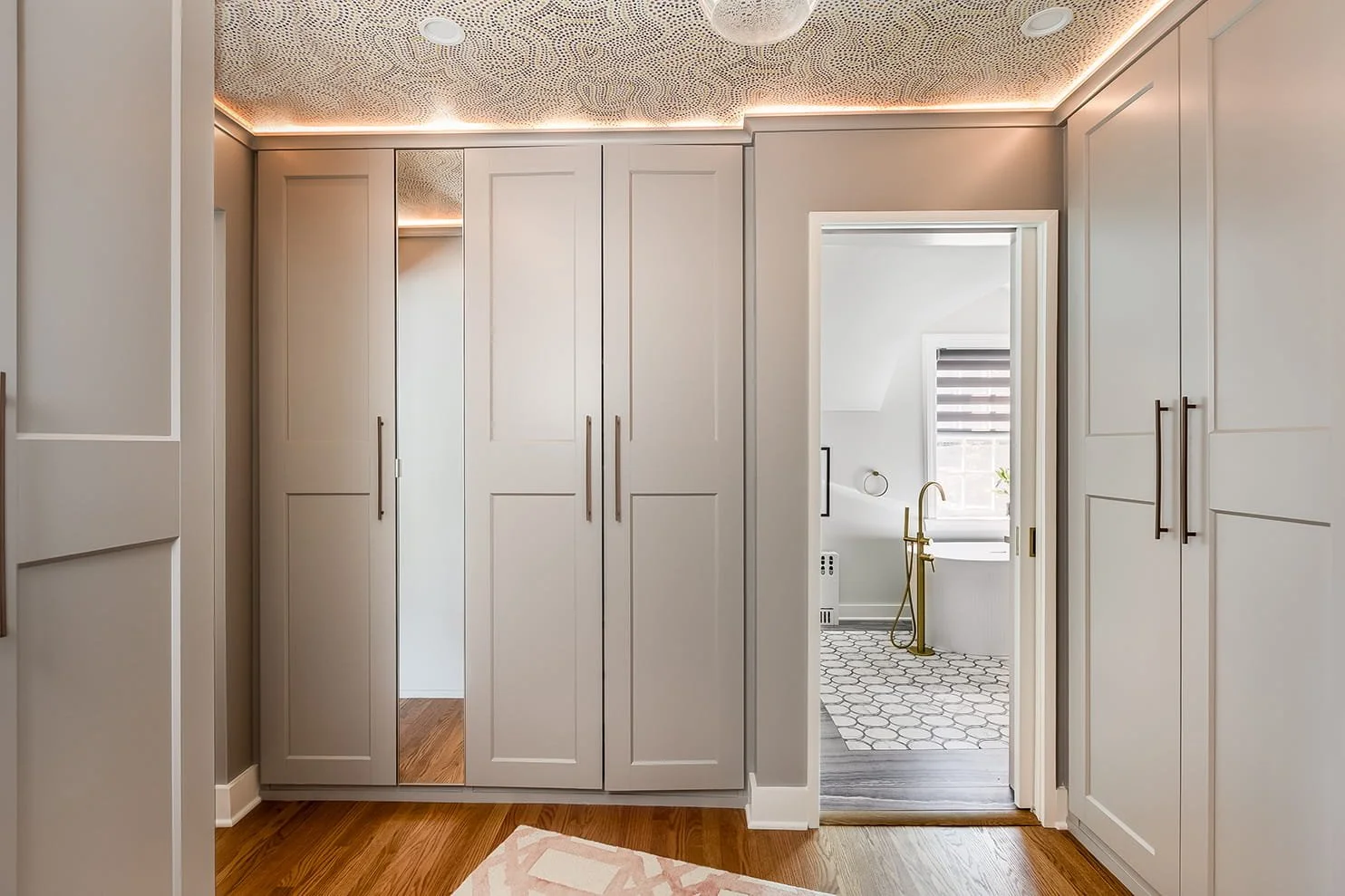 View of a walk-in closet with white cabinets and a doorway leading to a bathroom with patterned tile flooring, a bathtub, and a window with blinds.