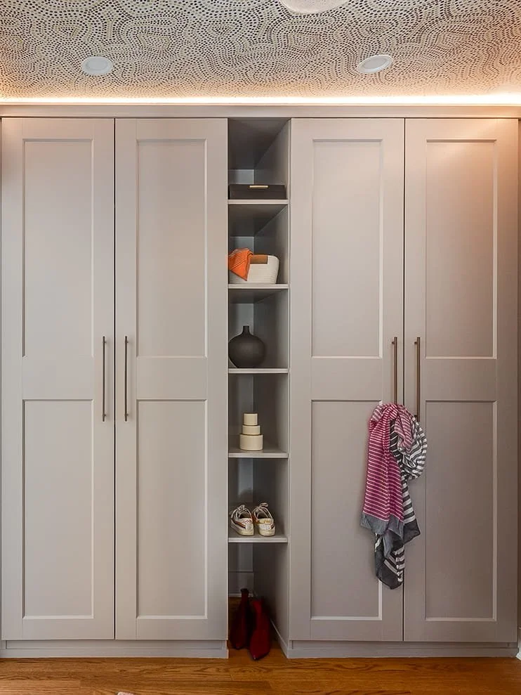 A white built-in closet with four large doors, brass handles, and a narrow open shelf column in the middle displaying decorative items, clothing, and shoes, with a towel hanging from one of the doors and a pair of red boots on the floor.