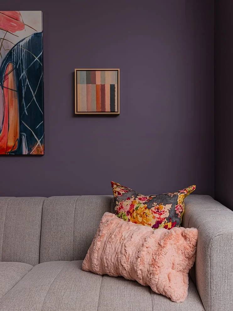 A gray sofa with two decorative pillows in front of a purple wall, which has two framed abstract art pieces hanging on it.