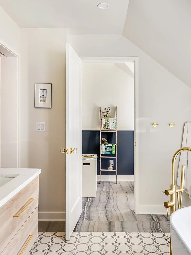 Bathroom with a vanity and gold fixtures, open door leading to a room with a ladder shelf and decorative items.