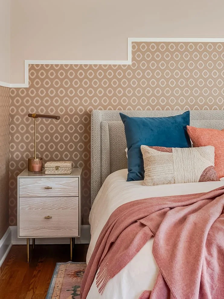 Bedroom with patterned beige wallpaper, white bedside table with gold accents, table lamp, decorative box, bed with blue, beige, and pink pillows, and a pink throw blanket.