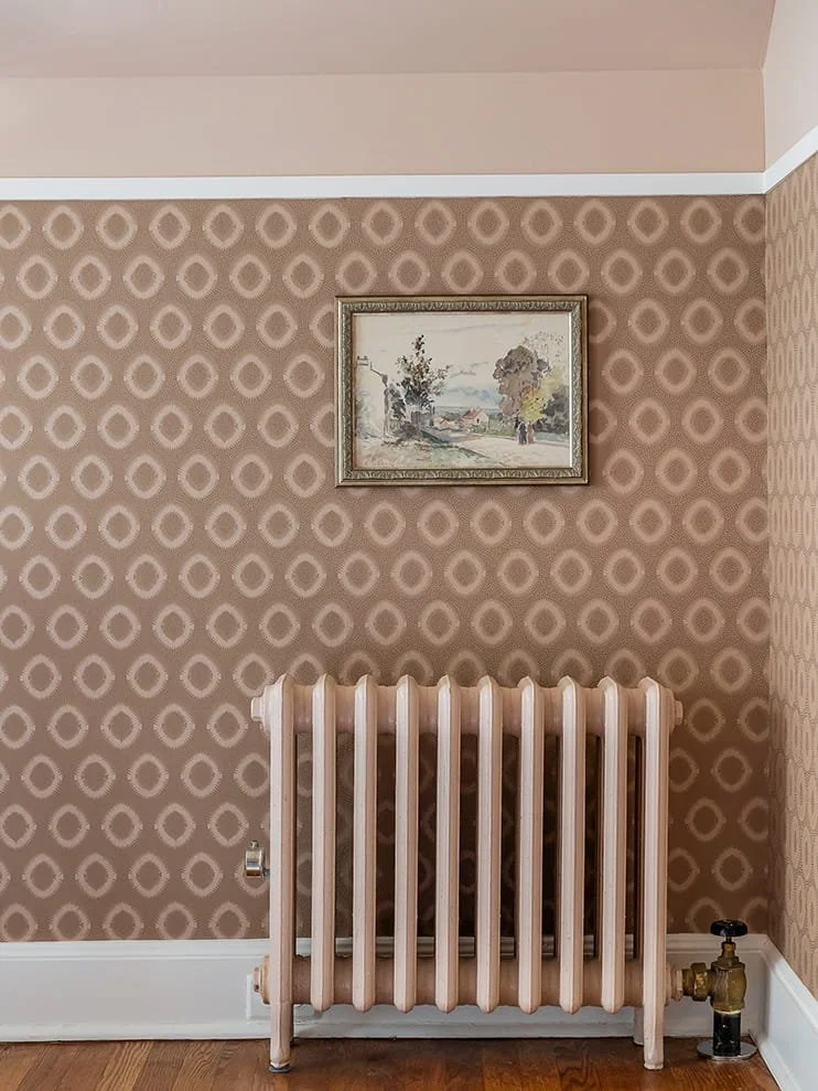 Interior scene with a vintage light pink radiator below a framed landscape painting on wallpapered wall with geometric pattern.