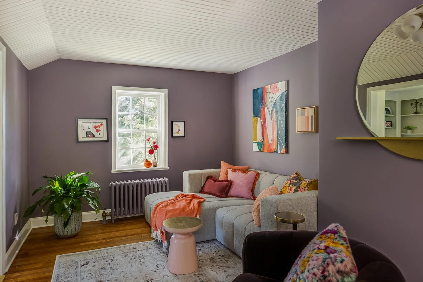 Living room with purple walls, white ceiling, hardwood floor, window with sunlight, sofa with pink and orange pillows, pink and orange blanket, colorful artwork on the wall, potted plant, round mirror, small side tables, and decorative items.