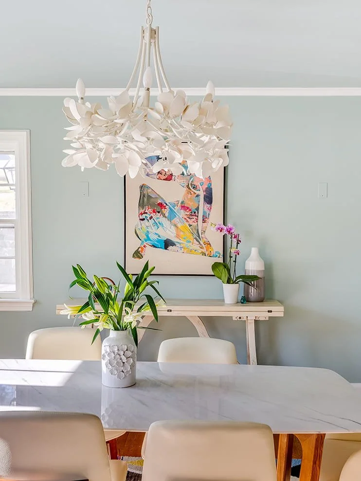 A dining room featuring a white marble table with a vase of white lilies, a light blue wall with an abstract colorful artwork, a wooden sideboard with a pink orchid in a white pot, a small white and pink vase, and a modern white chandelier hanging fr