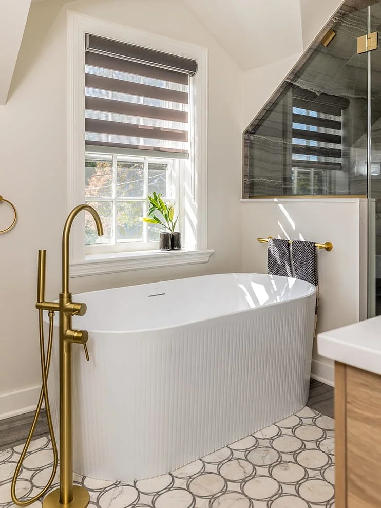 A modern bathroom featuring a freestanding white bathtub, a gold floor-mounted faucet with a handheld showerhead, a window with striped blackout shades, a small black planter with a green plant on the window sill, and a glass-enclosed shower with bla