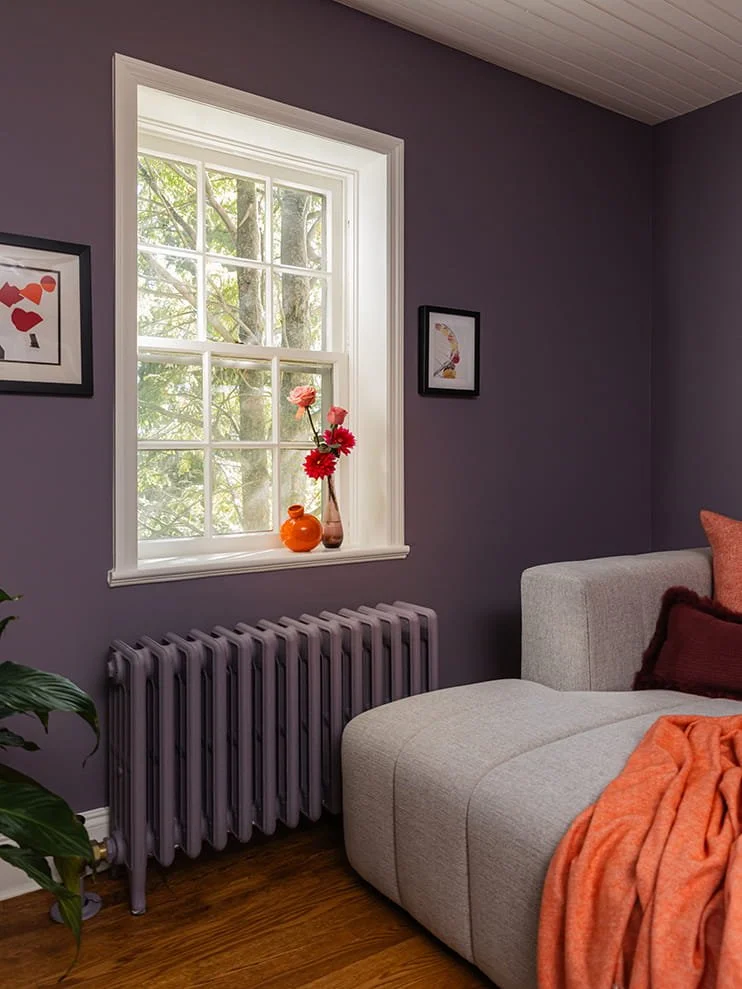 A cozy living room with purple walls, a window with white trim, a beige sectional sofa with pillows, a gray radiator below the window, and framed artwork on the walls. A vase with pink and red flowers is on the windowsill, with sunlight filtering thr