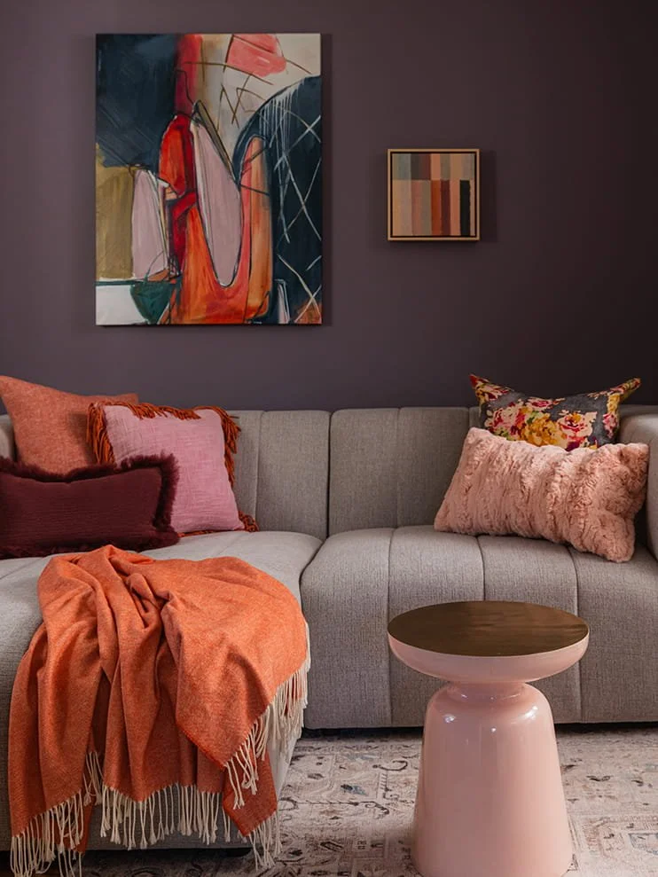Living room with a gray sectional sofa adorned with pink, orange, and floral pillows. An orange blanket is draped over the armrest. A small pink side table with a brown top stands in front. Dark purple painted walls feature two abstract paintings, on