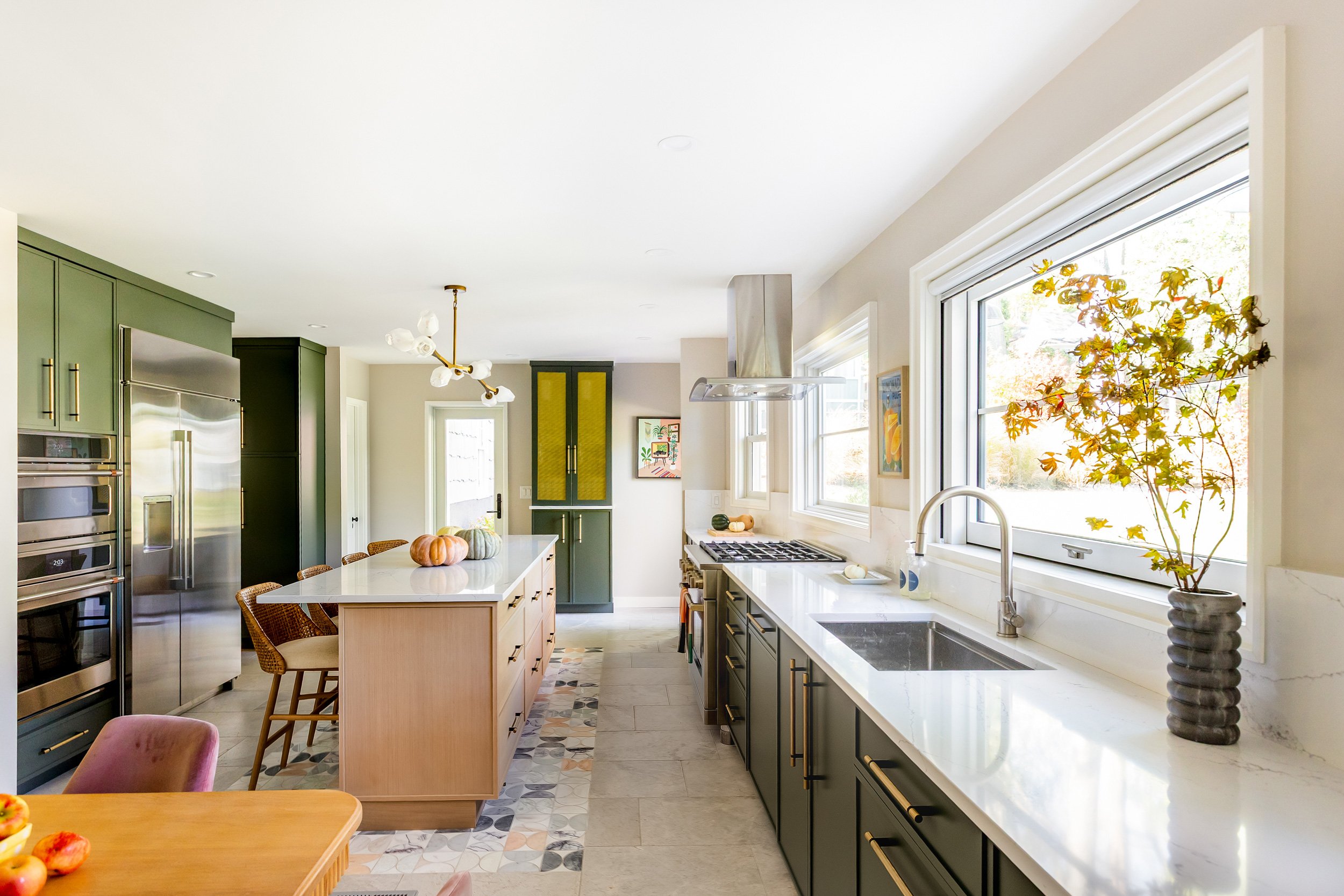A modern kitchen with green cabinets, a kitchen island, stainless steel appliances, and large windows with a view of outdoors, decorated with pumpkins and a potted plant.