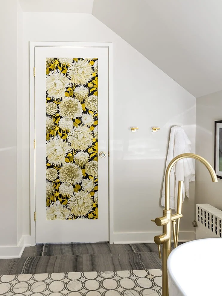 Bathroom with a door decorated with floral wallpaper, gold fixtures, white walls, a towel hanging on a hook, part of a bathtub with a gold faucet, and patterned floor tiles.