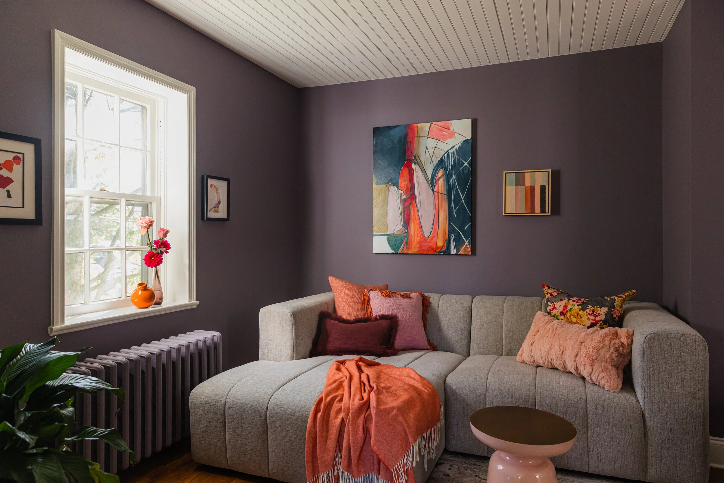 Living room with purple walls, beige sofa with pink, brown, and floral pillows, orange blanket, and small pink table. Window with white trim, vase of pink flowers on sill. Several framed abstract art pieces on wall.