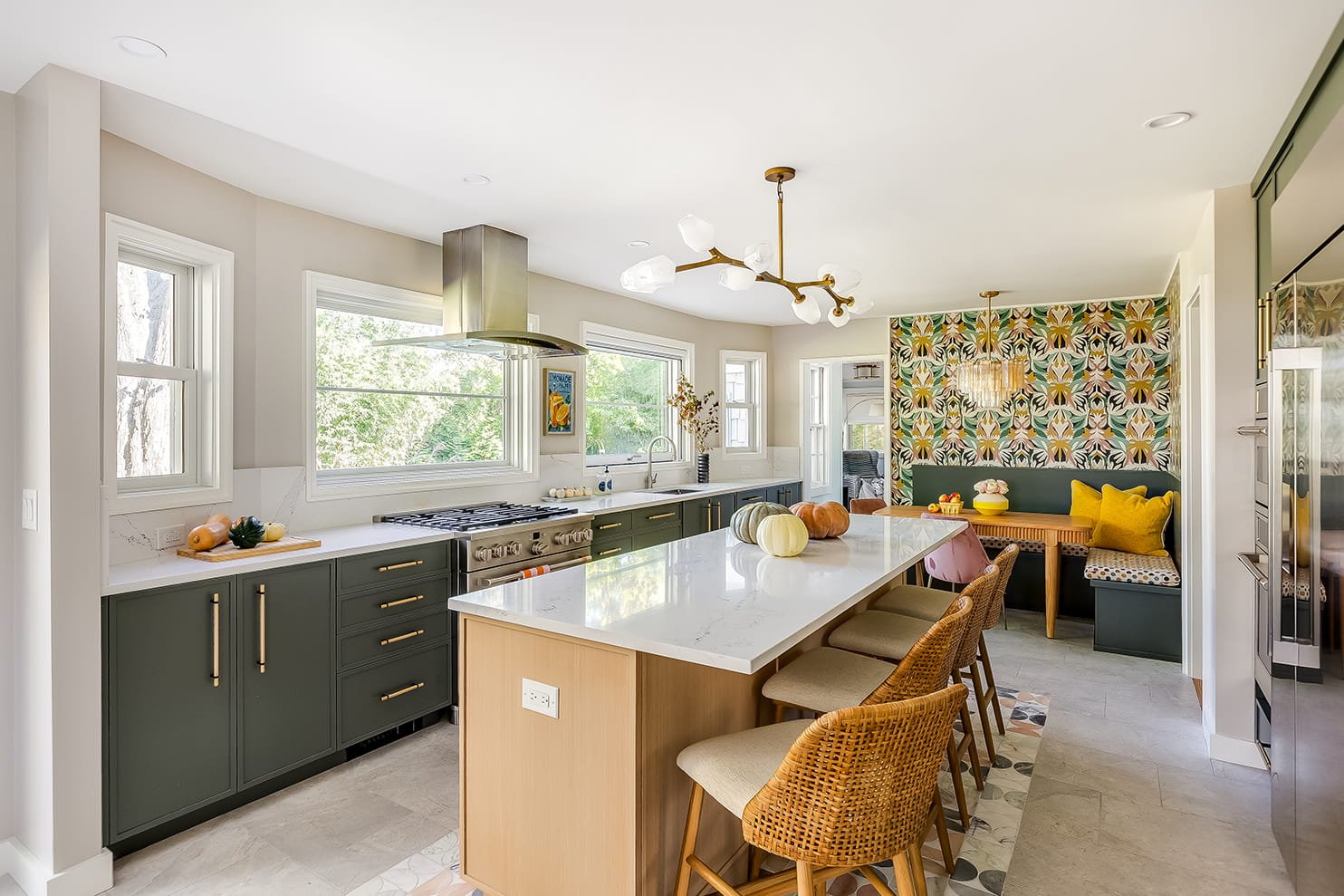 Modern kitchen with green lower cabinets, white countertops, large island with seating, colorful pumpkins on island, patterned wallpapered nook with bench, yellow and pink cushions, window overlooking trees.