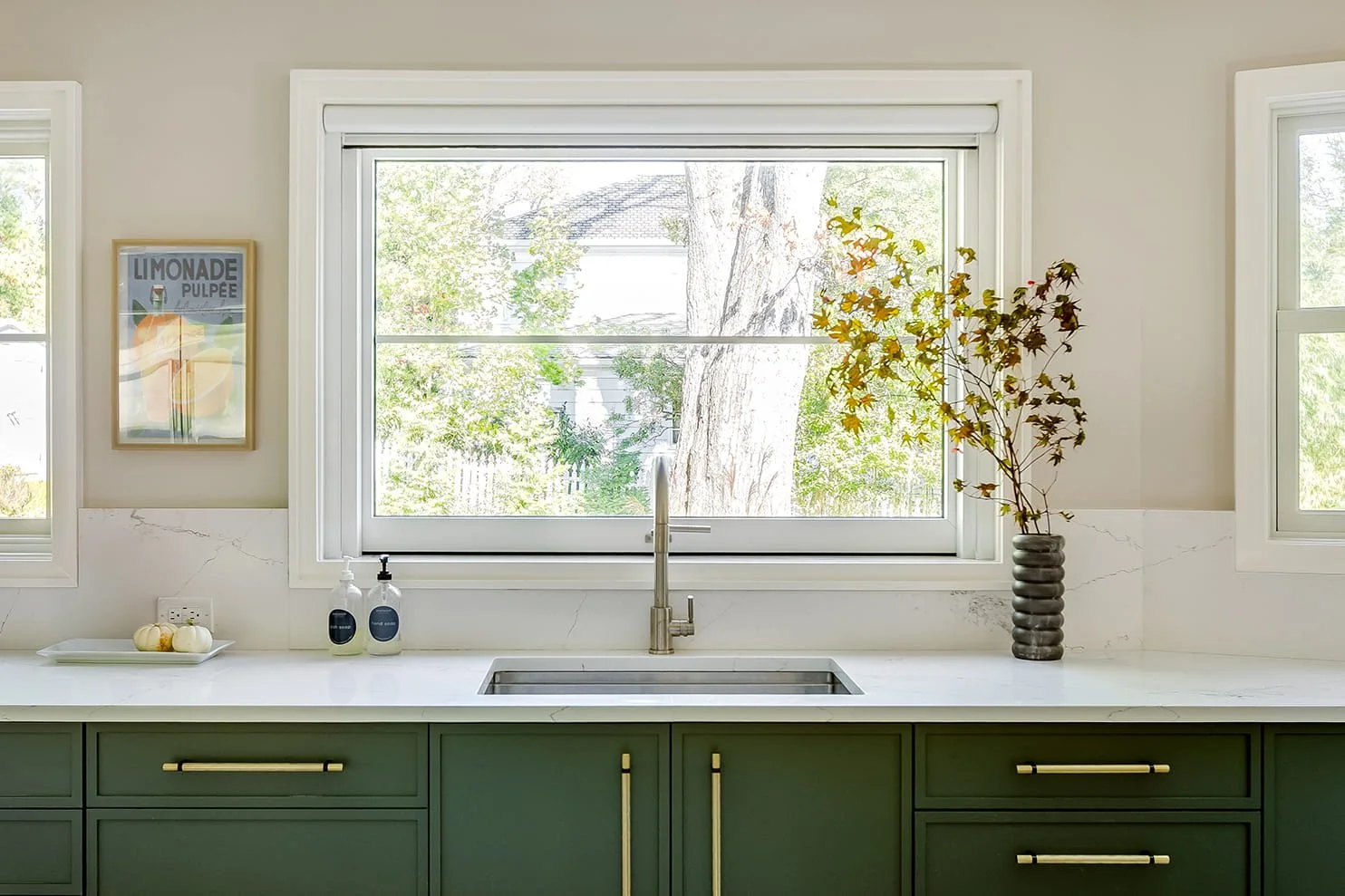 Kitchen window with view of trees, green cabinets, white marble countertop, sink, a vase with branches, two soap dispensers, and a framed print on the wall.