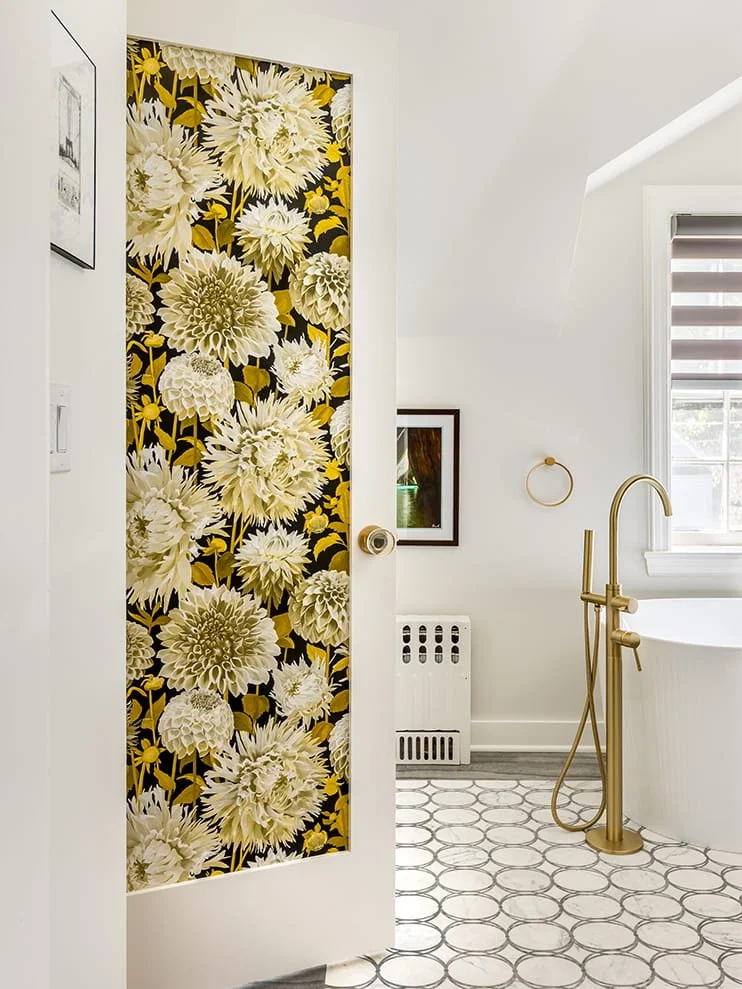Bathroom with a door featuring floral wallpaper, a bathtub with a gold faucet, a window with striped blinds, and a white wall with framed artwork.