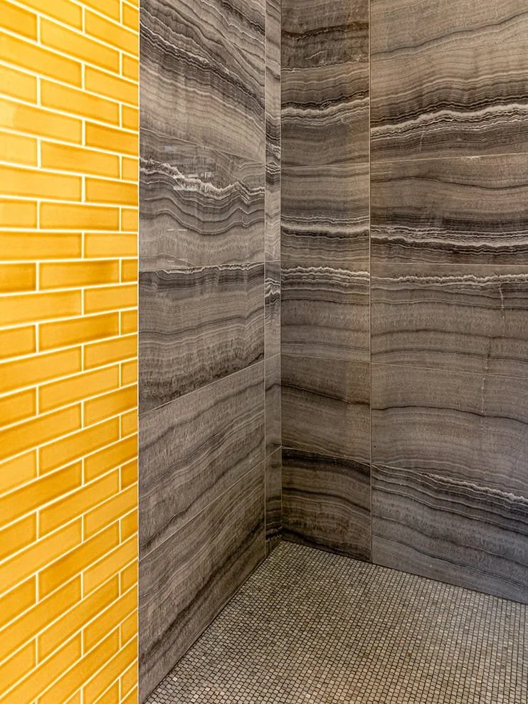 Corner of a room with a yellow brick wall on the left, gray marble or tile wall on the right and back, and small square mosaic tiles on the floor.