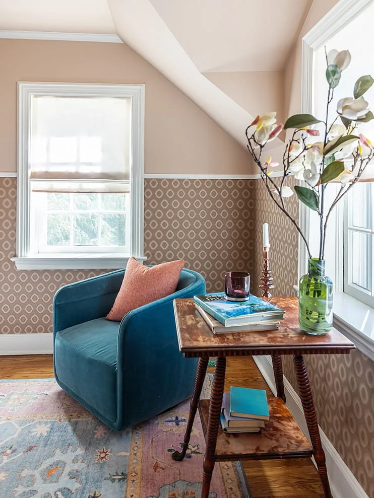 A cozy corner with a blue velvet armchair, a pink throw pillow, a wooden side table with books, a green glass vase with flowers, and two windows with white trim letting in natural light.