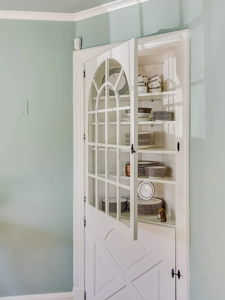An open white display cabinet with glass-paneled doors revealing stacked plates, bowls, and cups inside, situated against a pale green wall.