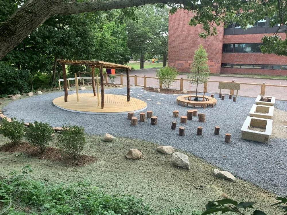 A view from the back of the Quincy Outdoor Classroom with new plantings, a new ground surface, seating areas, and a covered platform.