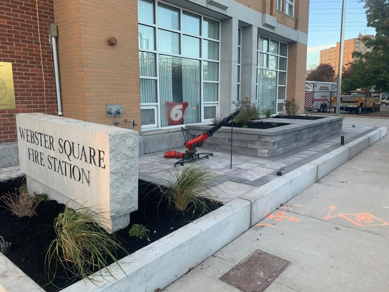 The granite sign for Worcester's Fire Fighter Memorial has new mulch around it and new plantings, and beside it is a statue of a hose.