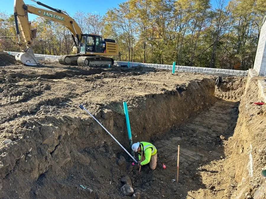 A worker kneels in a trench installing drainage for a retaining wall that is being constructed.