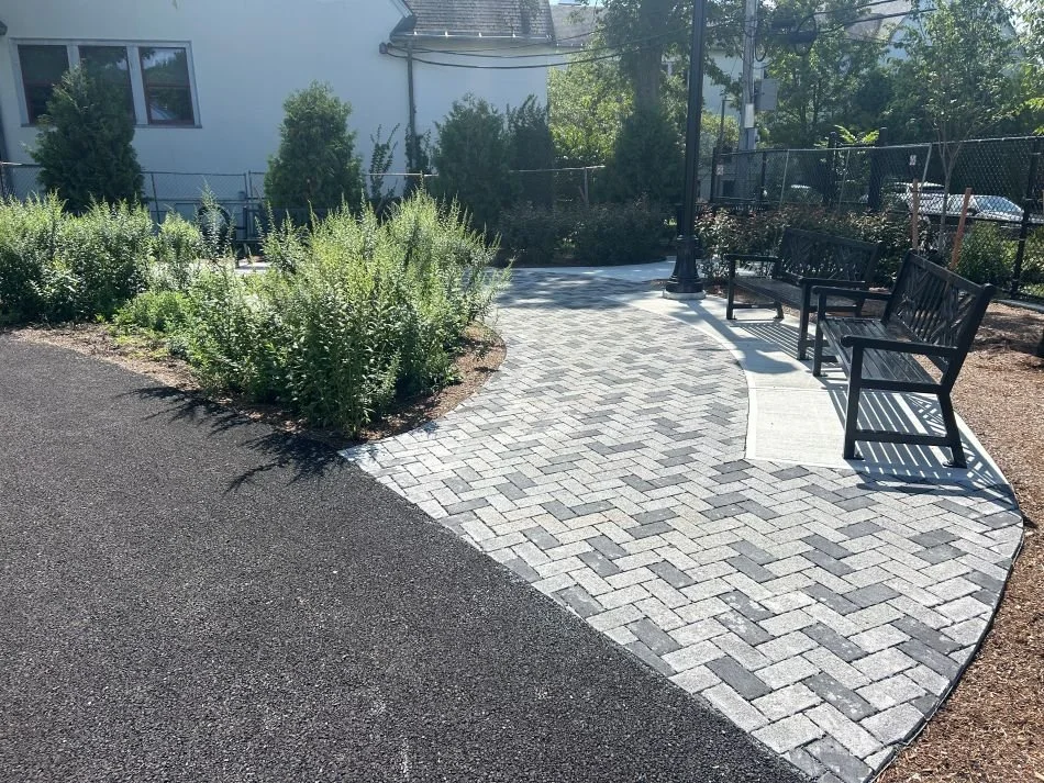 Dark and light pavers in a herringbone pattern make a U-shaped walkway and sitting area with benches around a mulched planting bed with  plants.