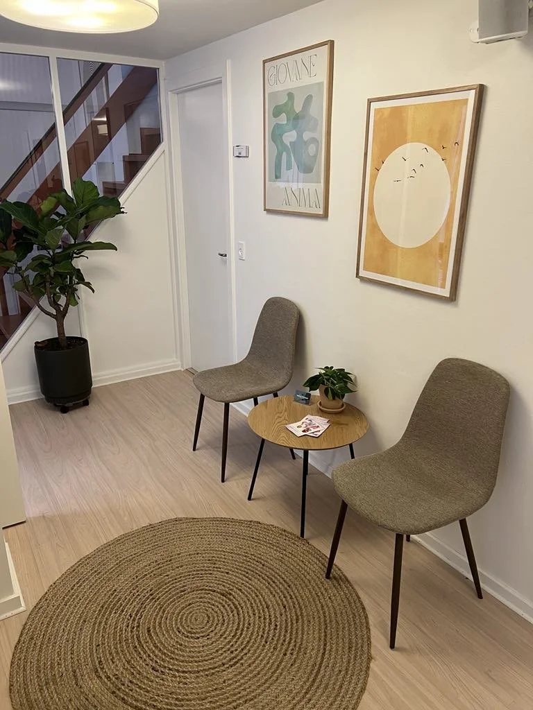 A cozy waiting area with two beige chairs, a small round wooden table with a potted plant, a magazine, and some cards.
