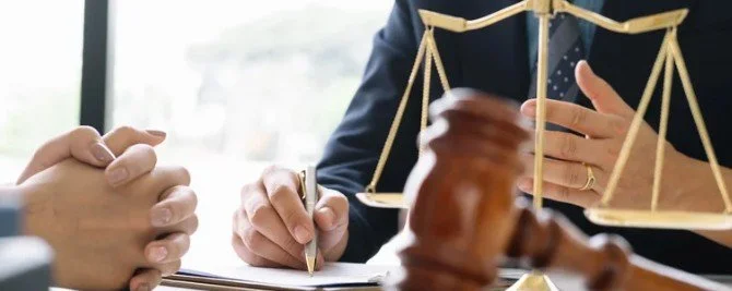 A person in a legal suit holding a scale of justice during a consultation with a client, with a gavel and legal documents on the table.
