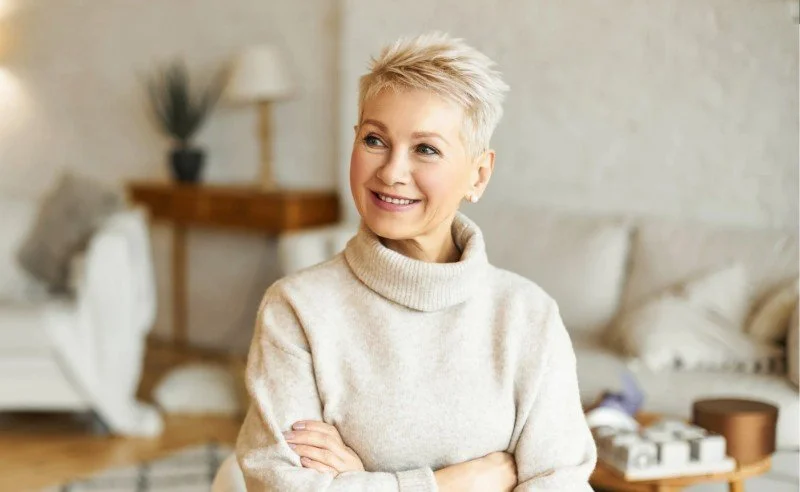 A woman with short blonde hair smiling and looking to the side, sitting in a cozy living room with beige walls and minimal decor.