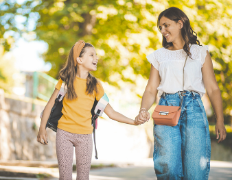 A woman and a girl walking outside hand in hand in a park on a sunny day.