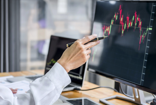 Person pointing at a financial chart on a computer monitor, with a laptop and tablets on the desk.
