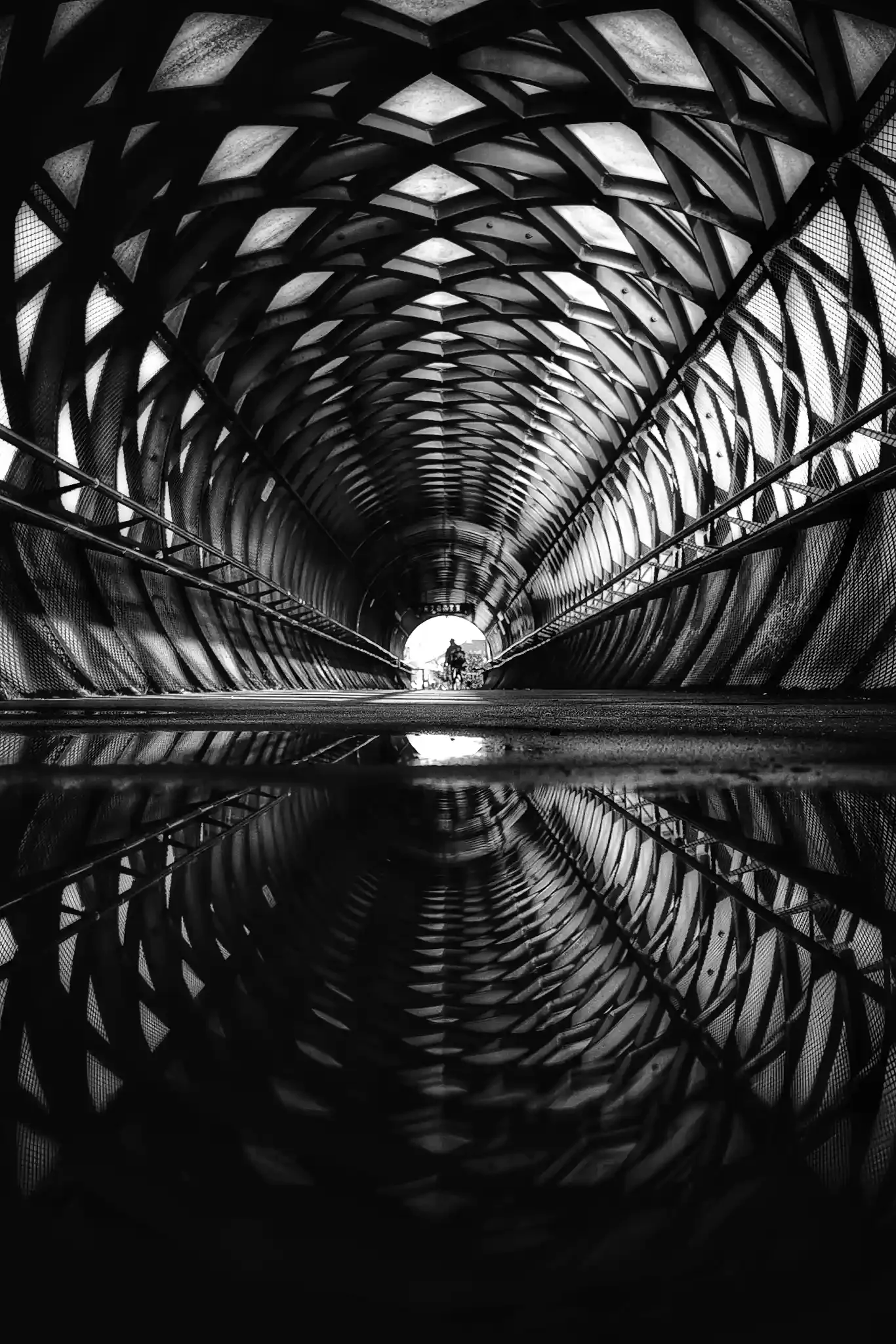 Passerelle Eiffel de La Roche-sur-Yon en noir et blanc, structure métallique en résille photographiée en symétrie avec reflet dans une flaque, une silhouette au loin dans la lumière