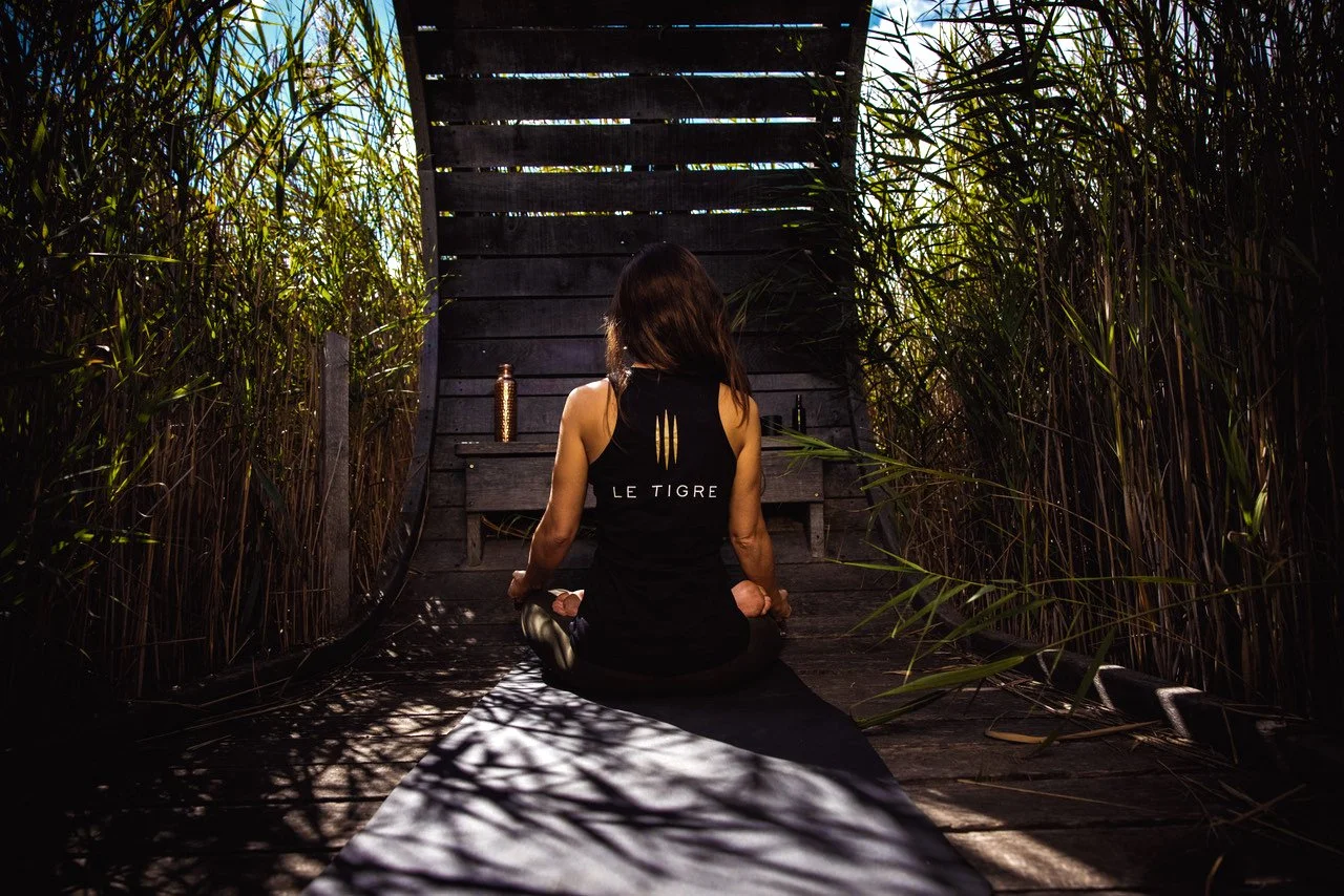 Femme faisant du yoga dans un environnement naturel entouré de végétation, assise sur un tapis de yoga, vue de dos.