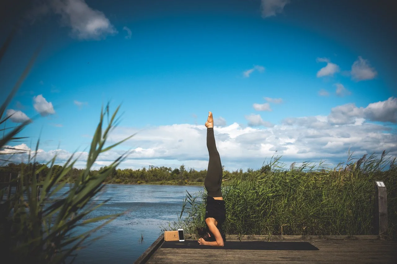Femme faisant une posture de yoga en équilibre sur la tête sur une plateforme en bois près d'une rivière, avec un ciel bleu et des nuages, entourée de hautes herbes vertes.