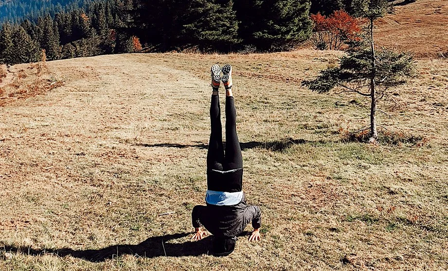 Une personne faisant une posture de yoga tête en bas dans un champ avec des arbres en arrière-plan.