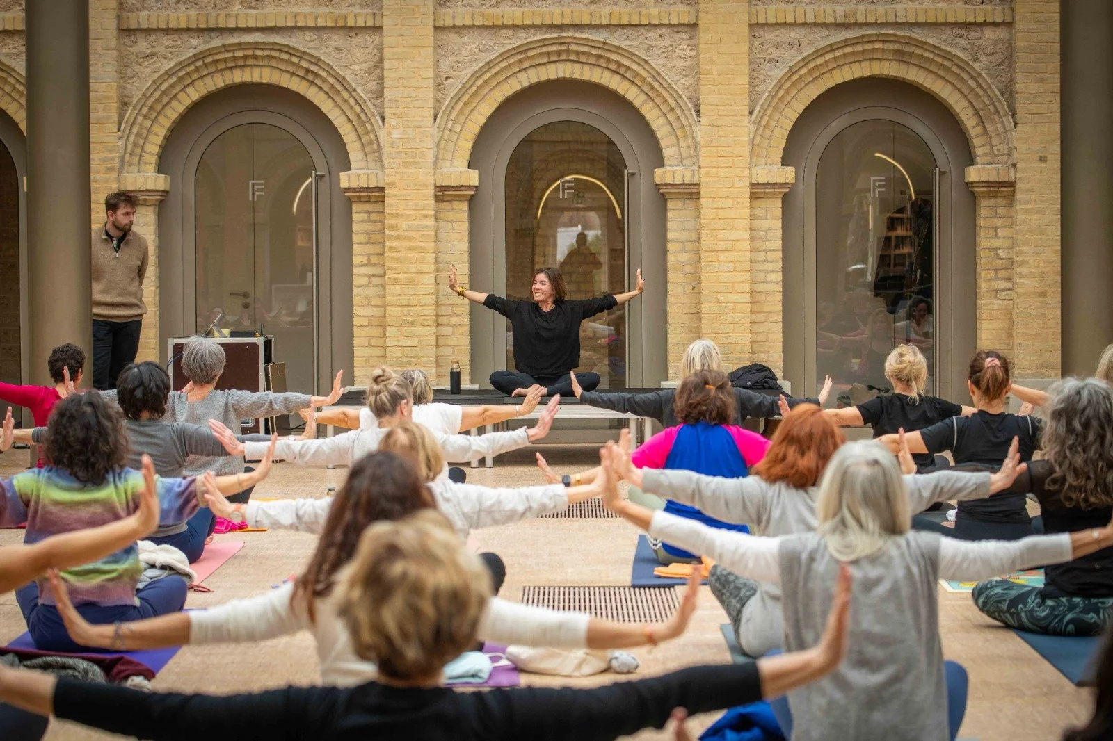 Groupe de personnes pratiquant le yoga en intérieur, assises sur des tapis, dirigé par Elodie Garamond assise en position de méditation avec les bras étendus, dans un espace avec des murs en briques et de grandes fenêtres.