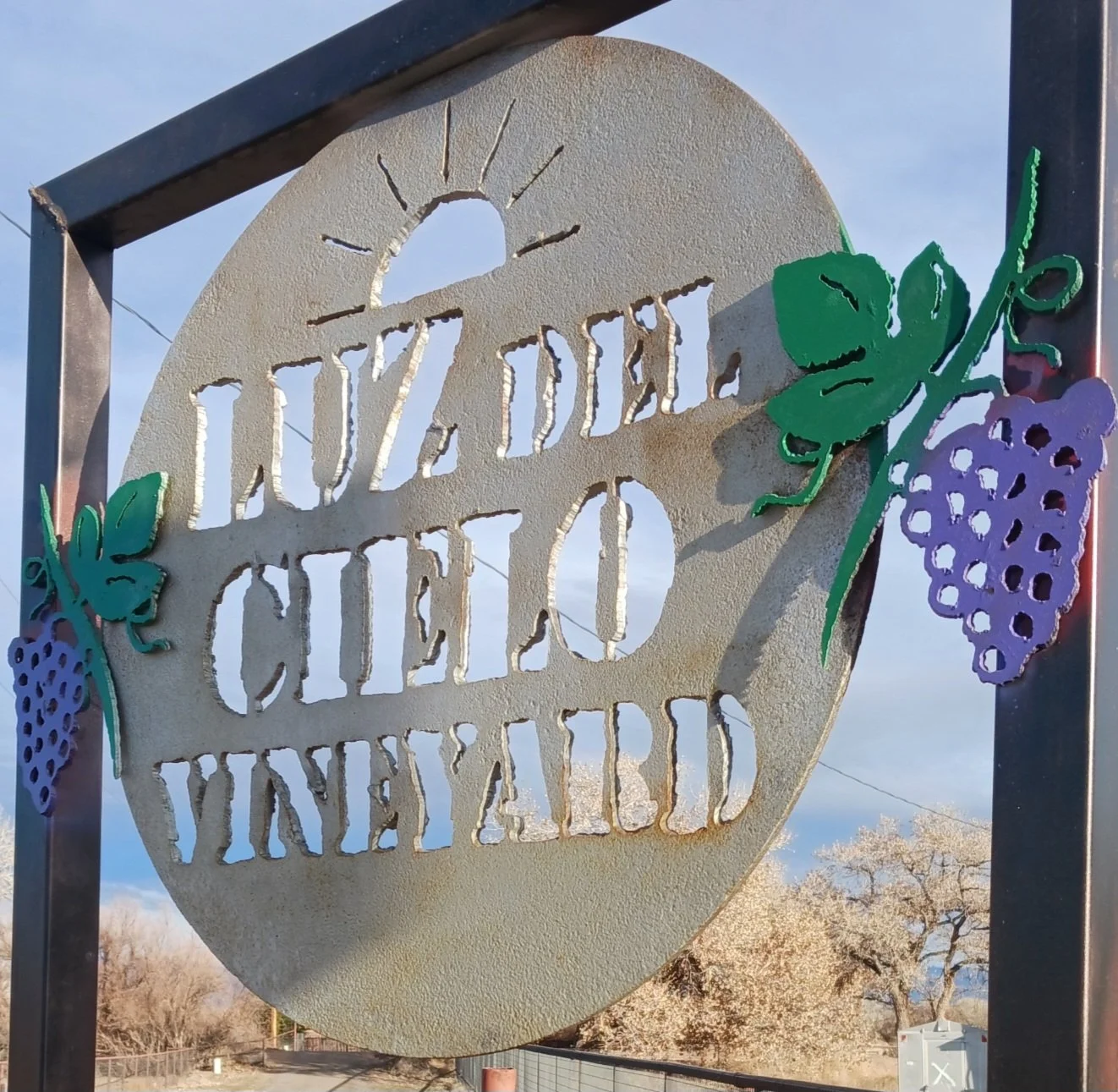 A decorative metal sign with the words 'Just Delicious Vine' cut out, decorated with purple grape clusters and green grape leaves on the sides.