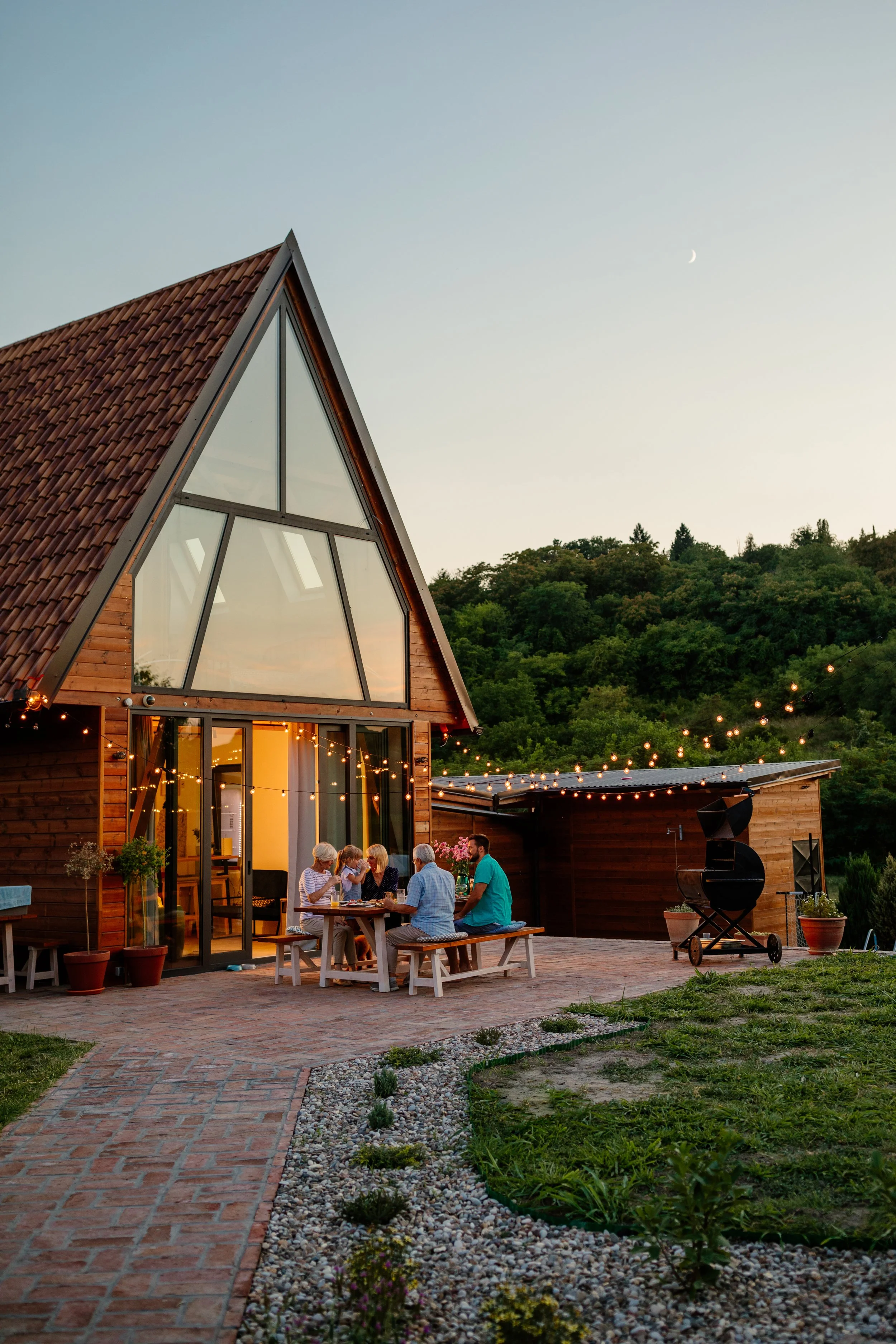 Family gathering at an outdoor patio of a modern wooden house during sunset, with string lights and a moon in the sky.