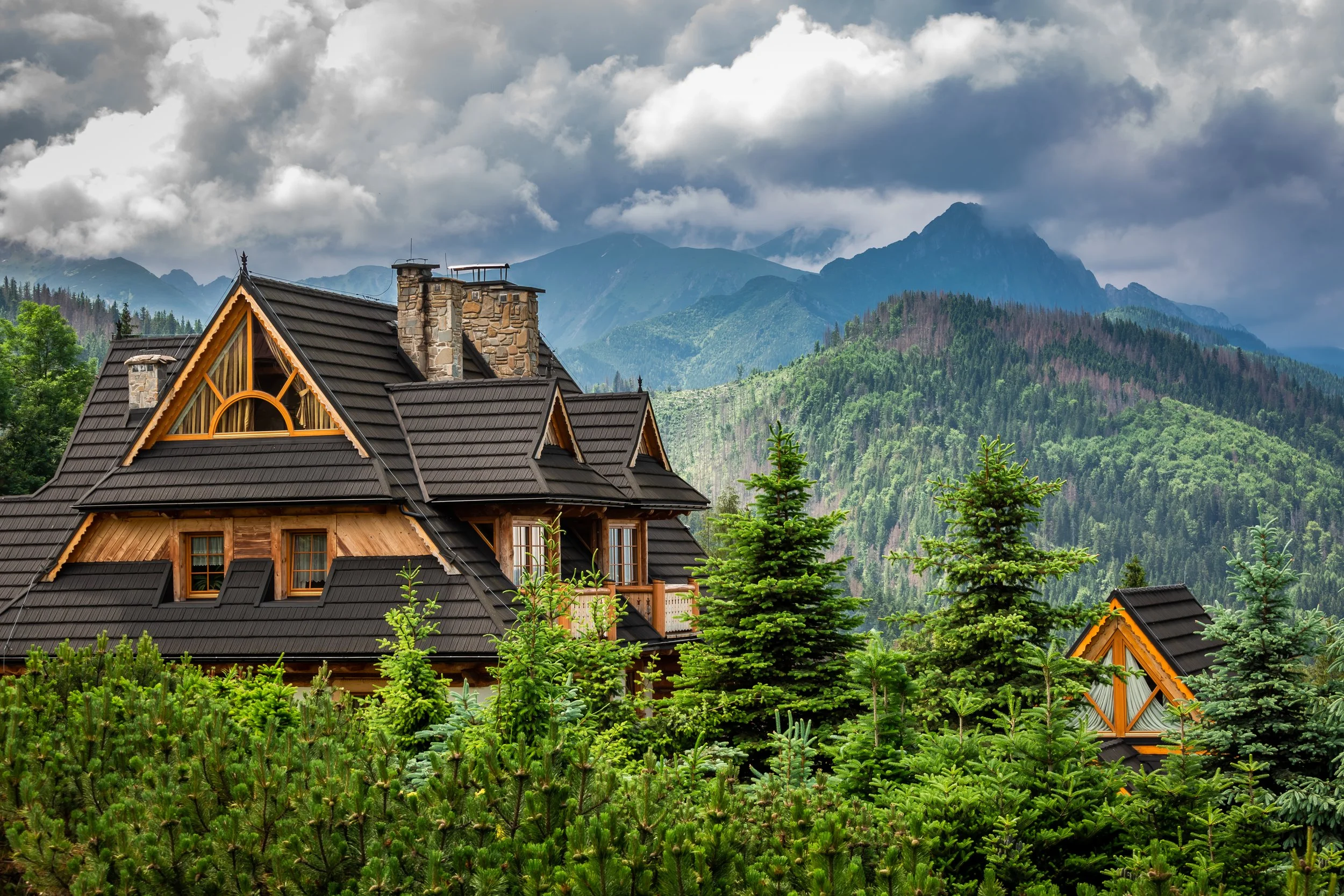 A wooden house with a dark roof on a lush green forested hillside, with mountains and cloudy sky in the background.