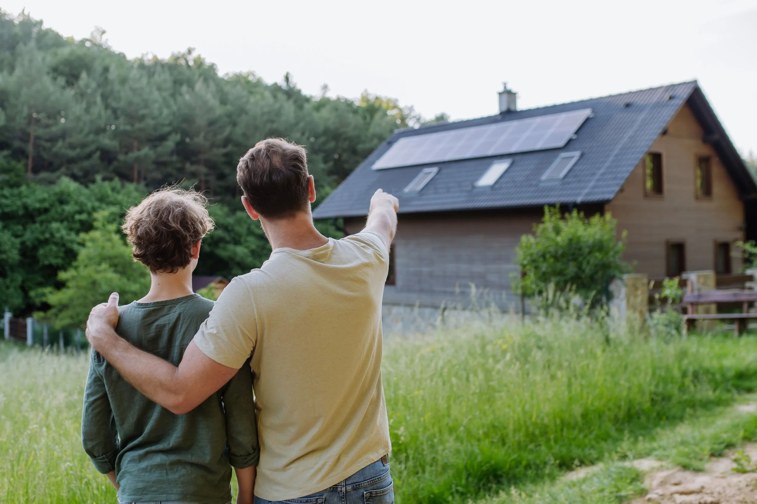 A man with his arm around a boy, pointing toward a house with solar panels on its roof, in a grassy area with trees in the background.