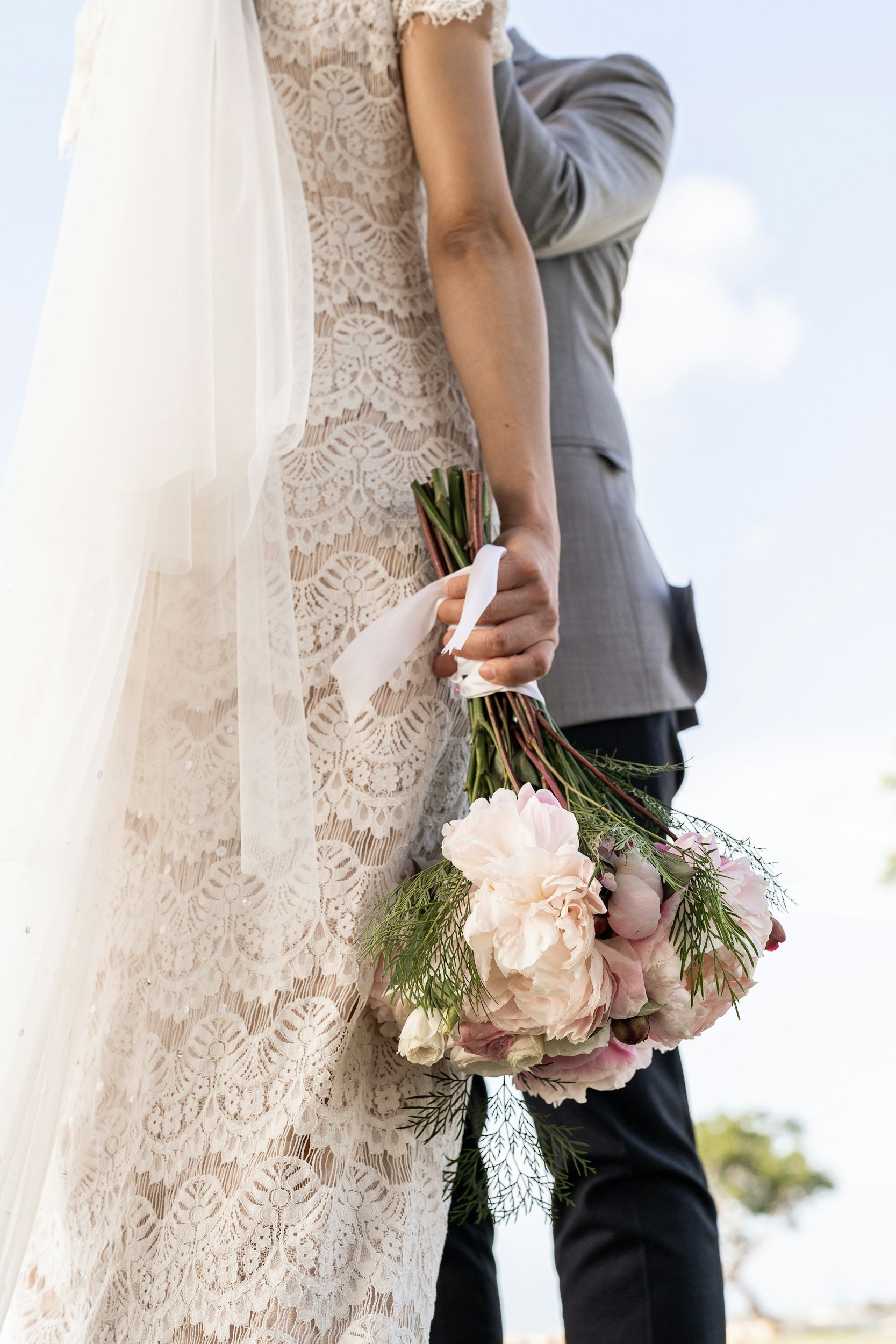A bride holding a bouquet of pink and white flowers, wearing a lace wedding dress, with a groom standing behind her in a gray suit.