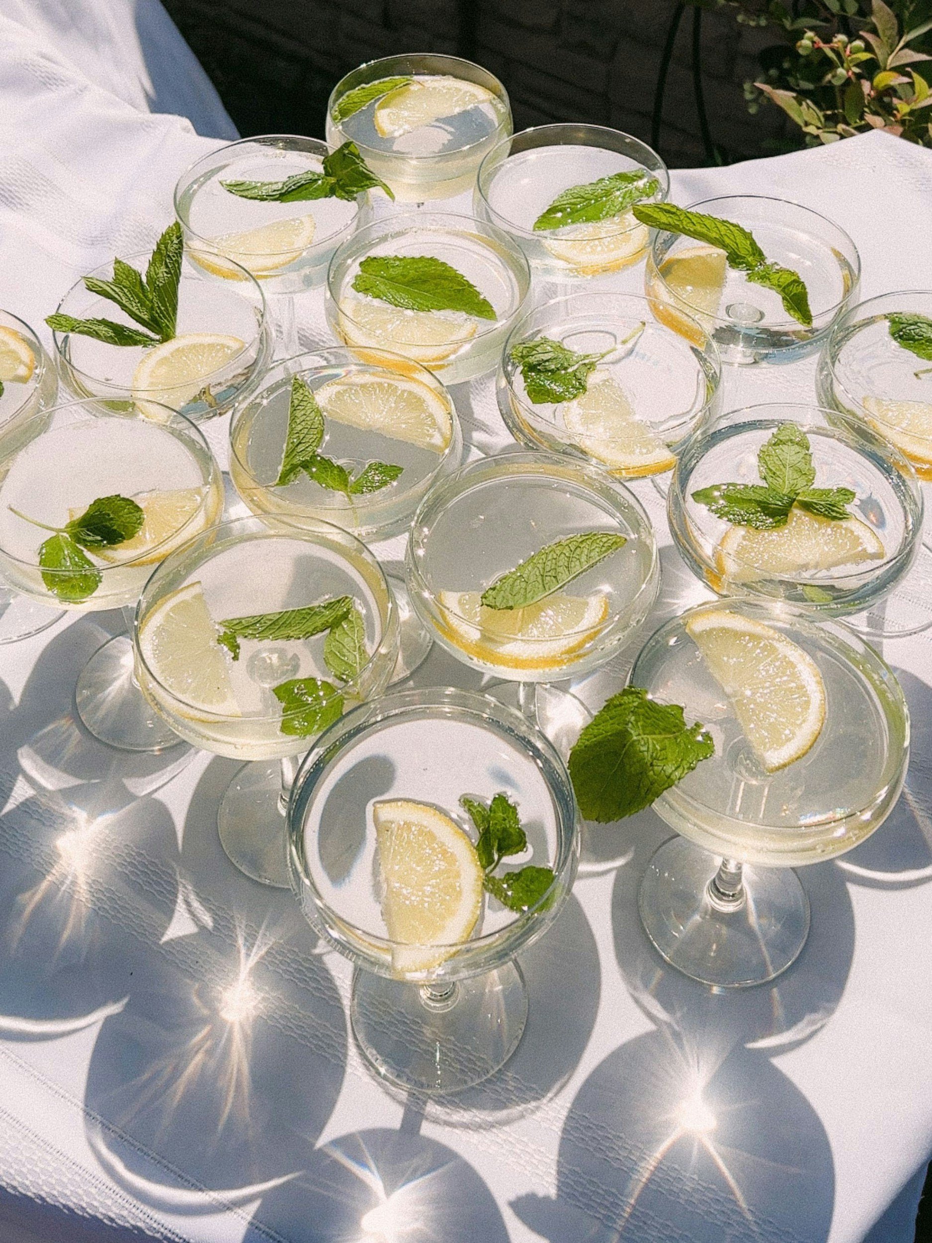 Multiple cocktail glasses with lemon slices and mint leaves on a white tablecloth.